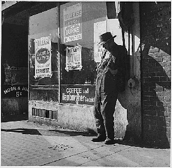 An unemployed and destitute man stands outside an empty store with a for lease sign.