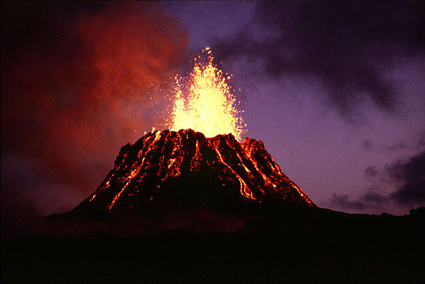 A nighttime photograph of a volcano cone with glowing lava flowing down the sides and lava bursting into the air