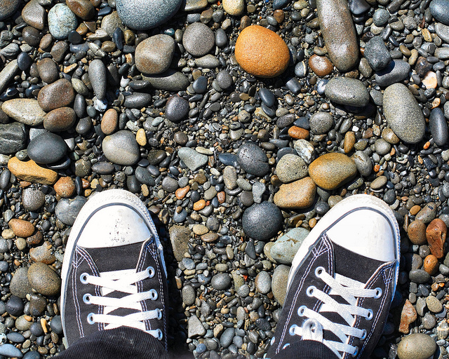 A point of view photograph looking down at the photographers sneakers standing on a gravelly beach.