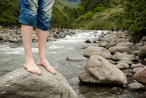 A photograph of a person’s bare feet standing on a rock by a stream.
