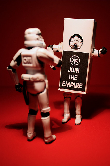 A photograph of a Star Wars action figure, a storm trooper looking at another storm trooper wearing a sign that reads “Join the empire.”