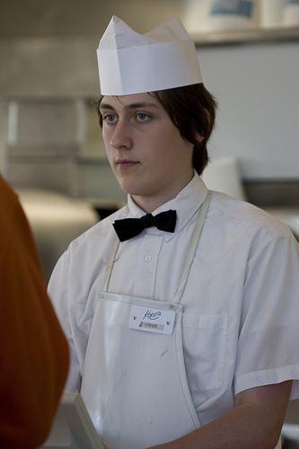 A photograph of a young man at work. He is wearing a uniform that consists of an apron, a bow tie, and a hat.”