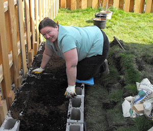A woman working on building a plant bed in her. She’s moved a row of cinder blocks into position. Her hands are covered with dirt, but she looks satisfied with her work.
