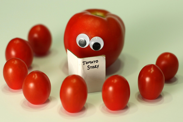 Still-life photo of a tomato with eyes glued on reading a story to smaller tomatoes