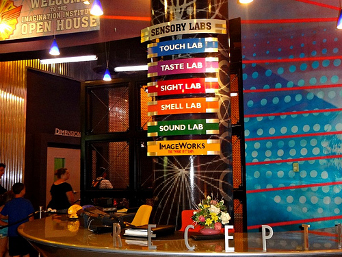 photo of a receptionist desk at a museum or art space. Signs that contain the words “Sensory Labs, Touch Lab, Taste Lab, Sight Lab, Smell Lab, Sound Lab, and Image works” hang above a fancy, crescent-shaped desk
