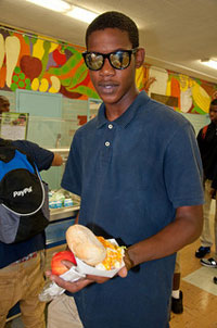  A photograph of a student holding his lunch out in front of him. He is standing in a school cafeteria.