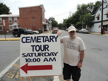 Photo of a man standing next to a sign promoting a “cemetary” tour; “cemetery” is misspelled.