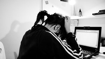 A photograph of a young woman holding her head, perhaps in frustration, while looking at a laptop computer screen