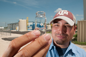 A man wearing a Texas A&M baseball cap holds up a small sample of water; an industrial plant is in the background.