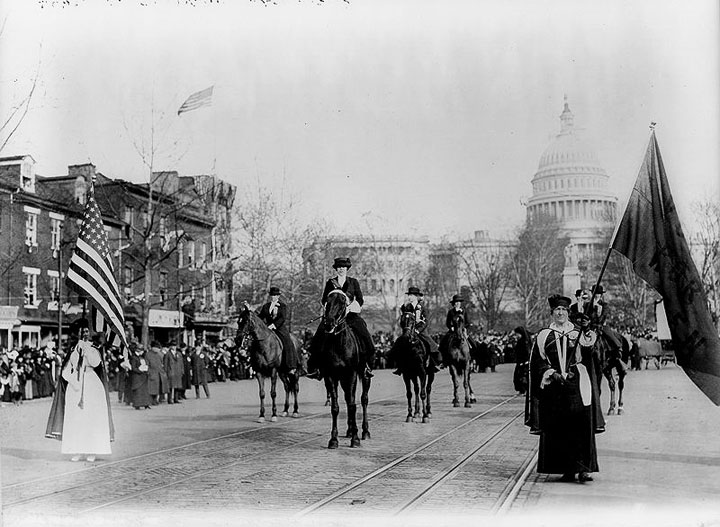 Black and white photograph (author unknown) of a suffragette parade.In the foreground are women dressed in black gowns with regalia. Several are on horses. In the background the US Capitol looms hazily. 