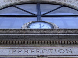 photo of a clock and beneath it the word “perfection” carved into the façade of an office building