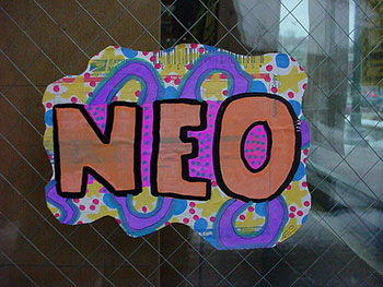 A photograph of the prefix “Neo” painted on cardboard and hanging on a fence.