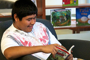 A photograph of a young man sitting in a library reading a graphic novel. He is smiling.