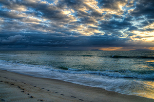 A photograph taken on a beach at dawn with clouds in the sky and footprints in the sand.