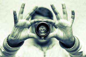 A photograph of a person looking through a detached camera lens. The image of the man is upside down.