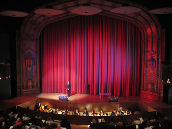 A photograph of a stage in a theater with an orchestra pit and an actress set off to the left.