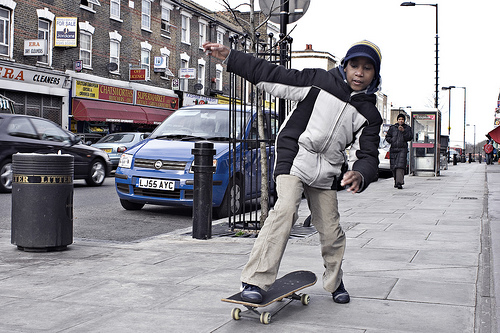 A photograph of a boy riding a skateboard on a city sidewalk. He is pushing off with his foot.