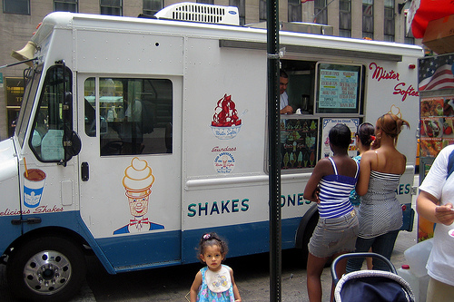 A photograph of an ice cream truck in a city. There are several young women and a little girl standing next to the truck.