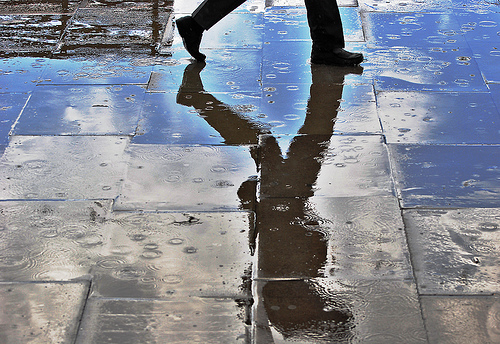 A photograph of a man walking on a sidewalk in the rain.