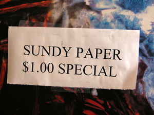 A printed sign in a store window that reads “Sundy Paper $1.00 Special.”