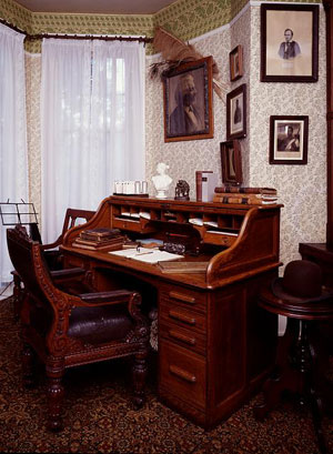 photo of Frederick Douglass den; shows an old roll top desk, and several photos hanging above the desk on a wall covered with antique wallpaper