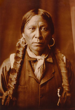 A photograph of an Apache man. He has long hair braids and is wearing hoop earrings.