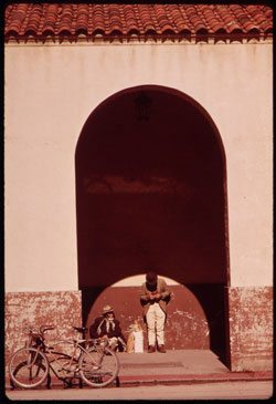 A photograph of the outside of a building in old San Antonio. There are two men talking under an awning and a bicycle propped up near them.