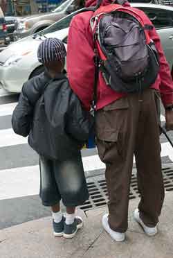 A photograph of a father and son waitng for a crossing light on a street corner. They are holding hands.