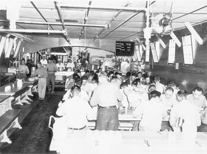 A photograph of the dining facility at a Japanese internment camp. There are a several tables full of people eating.