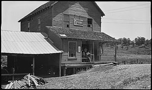 A photograph of an old saw mill. There are men sitting on the porch and a pile of cut lumber visible.