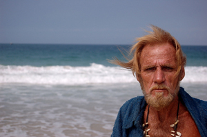 A photograph of an older man standing on a beach. He is bearded and weathered much like Robinson Crusoe must have looked.