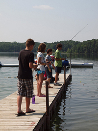 A photograph of several young people fishing from a pier, on a lake.