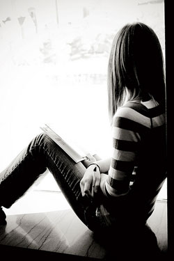 A photograph of a young woman, seated, looking out if a window. She is holding a notebook and a pen.