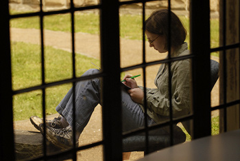 A photograph of a young woman taken through panes of glass in a window. She is writing in a notebook.