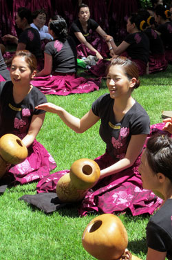 A photo of several young women all dressed in decorated t-shirts and pinkish/purple floral skirts as they sit in a grassy area and play drums shaped liked hourglasses