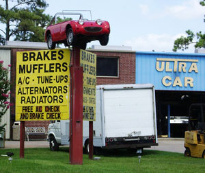 Photo of the front of a repair shop. A sign out front advertises services. on top of the sign is a small sports car.