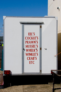 A fish vendors truck that reads “Eel’s, cockle’s, prawn’s, mussel’s, whelk’s, winkle’s, crab’s, etc.”