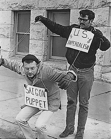 Two male protesters mimicking a puppet show with the man pulling the strings wearing a sign that indicates that he is 'US Imperialism'. The puppet is a man wearing a sign that indicates he is 'Saigon'.