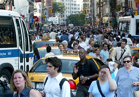 Image of a New York City street that is crowded with pedestrians. There several buses and taxi that are trying to make their way through the crowd.